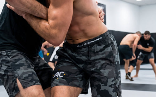 Two grapplers clinching on the mat in ALL CORNERS grayscale camo shorts, branded waistband visible